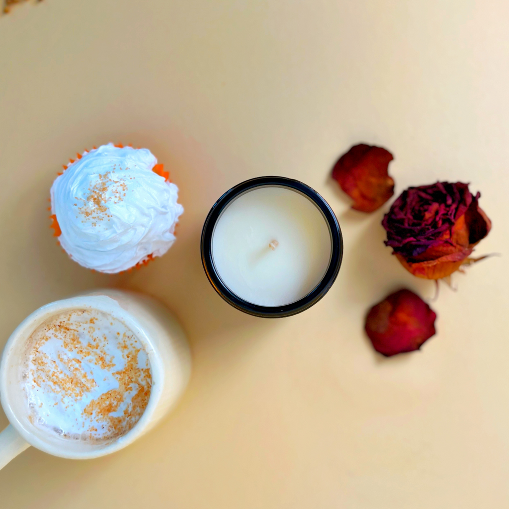Duftkerze Cozy Mood von Chris P. Candles auf einem Tisch, daneben liegen getrocknete Rosenblätter und ein Vanille-Muffin.