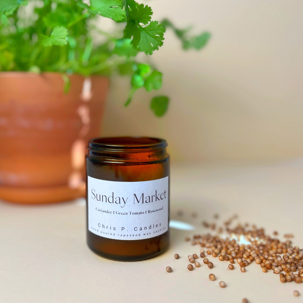 Glas-Duftkerze ‚Sunday Market‘ auf einem hellen Holztisch. Im Hintergrund ein Korainder-Pflanzentops. Die Kerze ist handgegossen in Berlin aus veganem Rapswachs. 